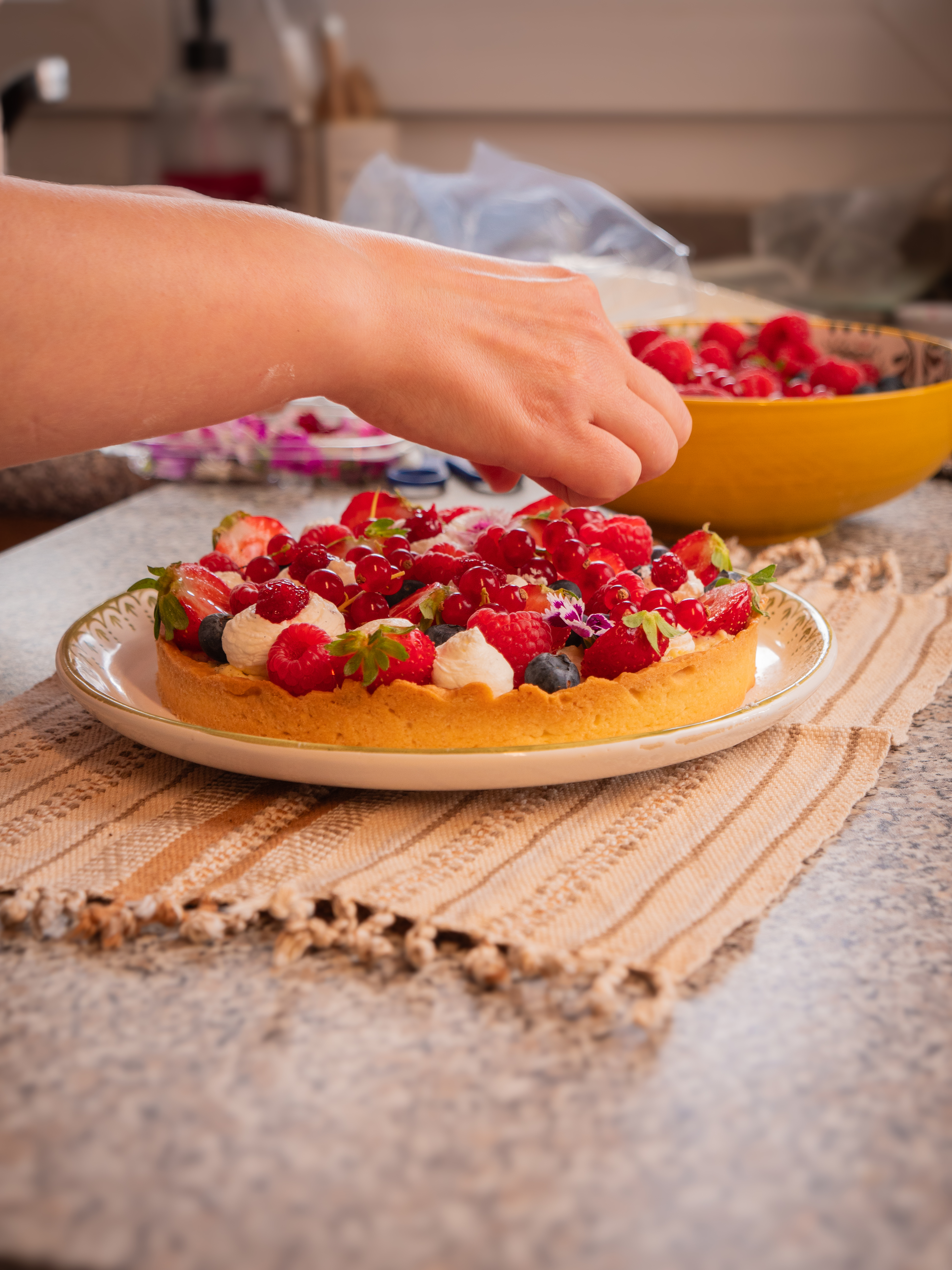 Une main dépose des framboises sur une tarte aux fruits rouges